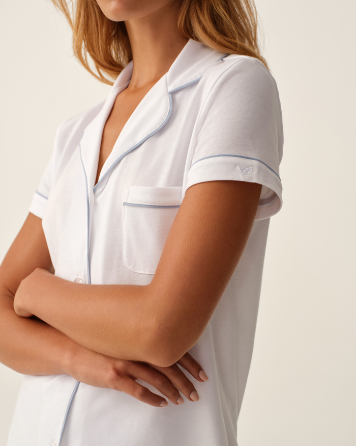 A woman with light brown hair stands with arms crossed, wearing the Greek Embroidered Poppy Pima Short Set in white, featuring periwinkle piping. Only her upper body is visible against a neutral background.