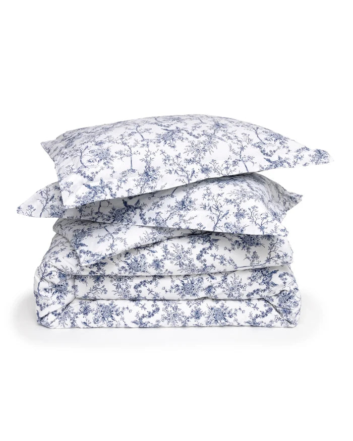 A neatly made bed features the Timeless Toile Bedding Set in white and blue with matching pillowcases, paired with a wooden headboard and a lamp on the nightstand in an elegant, well-lit bedroom.