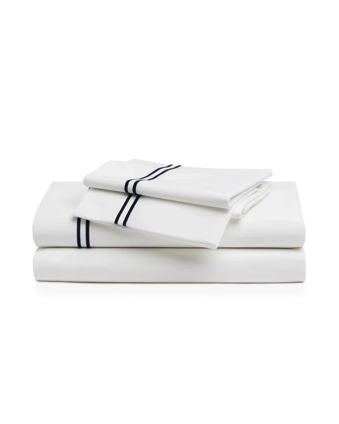 A neatly made bed features the Timeless Toile Bedding Set in white and blue with matching pillowcases, paired with a wooden headboard and a lamp on the nightstand in an elegant, well-lit bedroom.