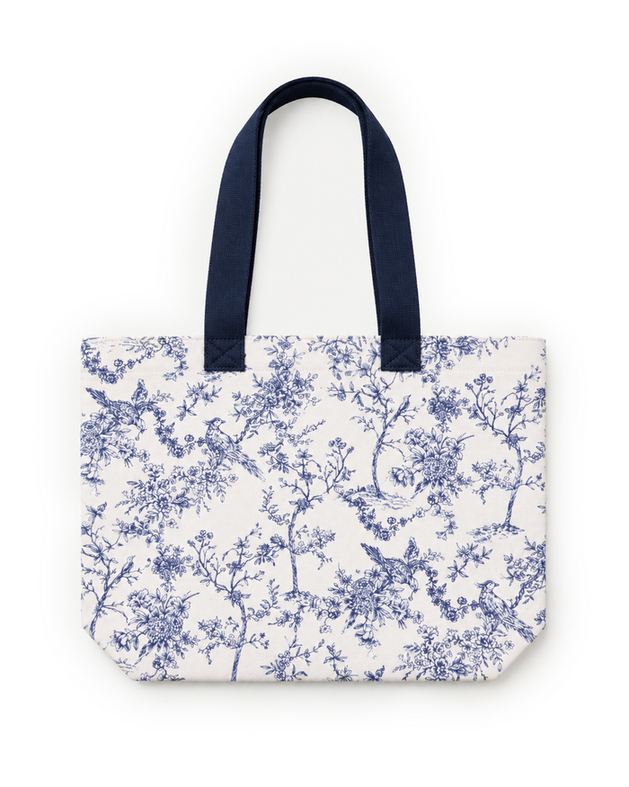 The Timeless Toile Tote Bag features blue handles and a blue toile pattern with birds, tree branches, and flowers. It is photographed against a white background.
