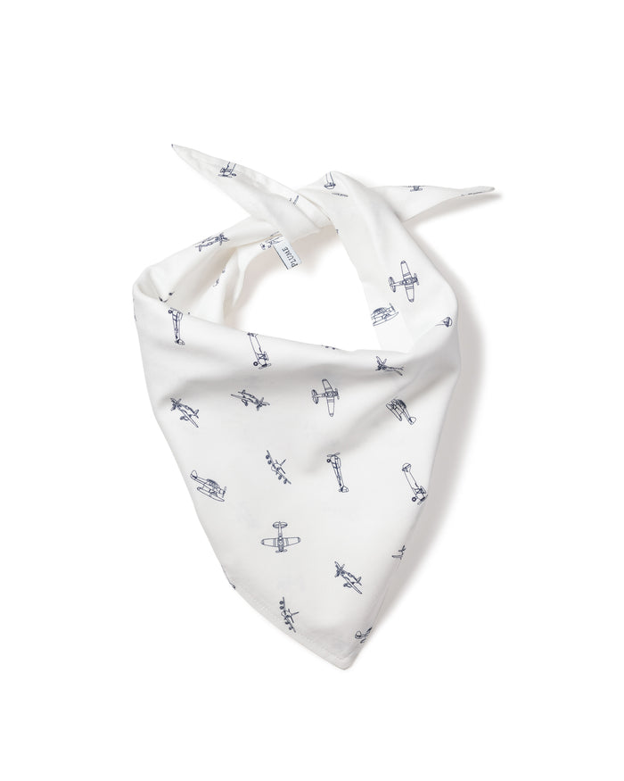 A fluffy white dog with small ears wears the Petite Plume Par Avion Dog Bandana featuring a blue floral pattern. The happy pup lies on a light wooden floor, looking up with its mouth open, showcasing the perfect blend of style and comfort in dog accessories.