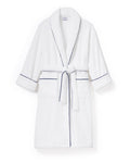 The Unisex Luxe Terry Robe in White with Navy Piping has a shawl collar, front pockets, waist tie, rolled sleeves, and dark piping along the edges.