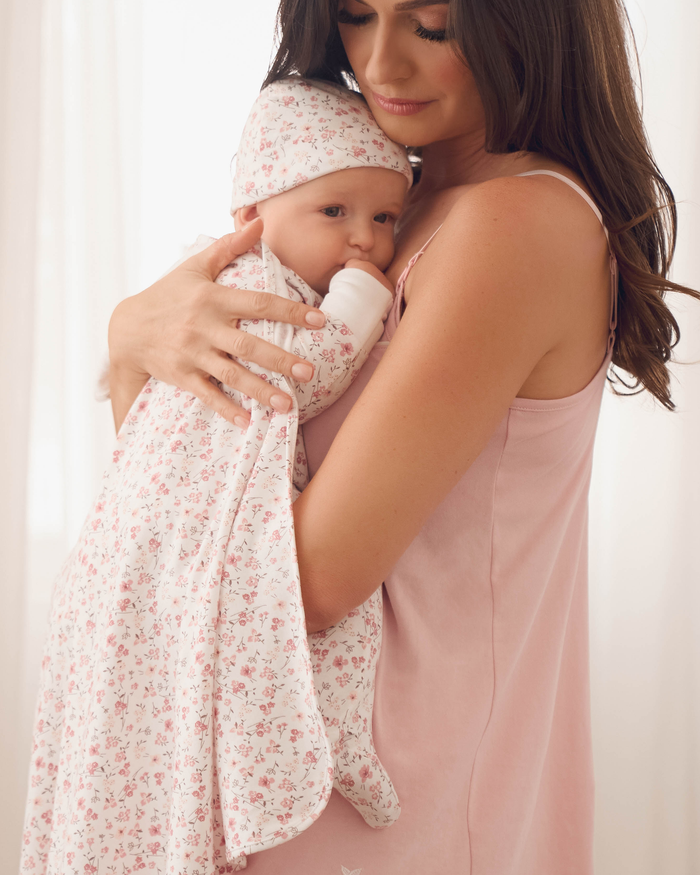 A woman in a pink dress lovingly holds a baby wrapped in Petite Plumes Welcome Home Baby Set - Dorset Floral. The baby, in an adorable romper, gazes at the camera against a softly blurred background.