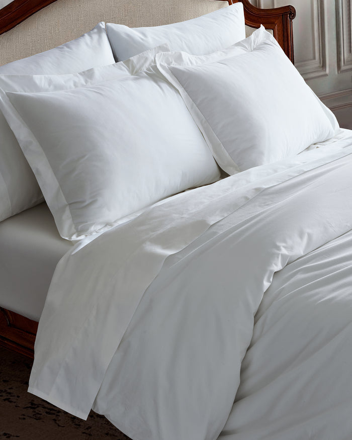 The White Bedding Set includes crisp white sheets, pillowcases, and a comforter, with multiple pillows arranged neatly against a light upholstered headboard in a softly lit bedroom.