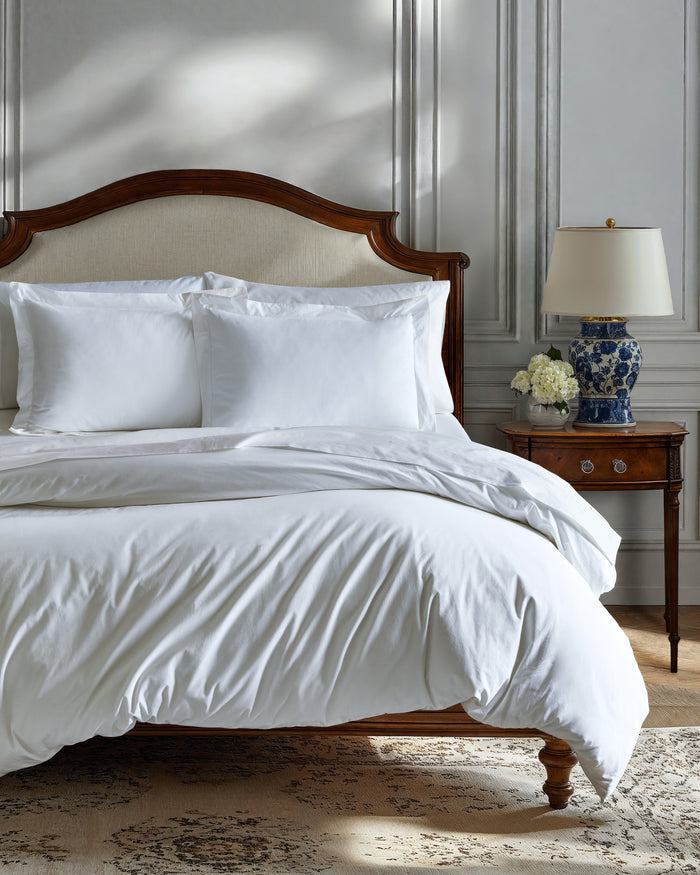 The White Bedding Set includes crisp white sheets, pillowcases, and a comforter, with multiple pillows arranged neatly against a light upholstered headboard in a softly lit bedroom.