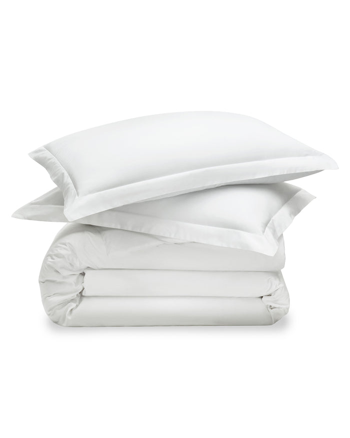 The White Bedding Set includes crisp white sheets, pillowcases, and a comforter, with multiple pillows arranged neatly against a light upholstered headboard in a softly lit bedroom.