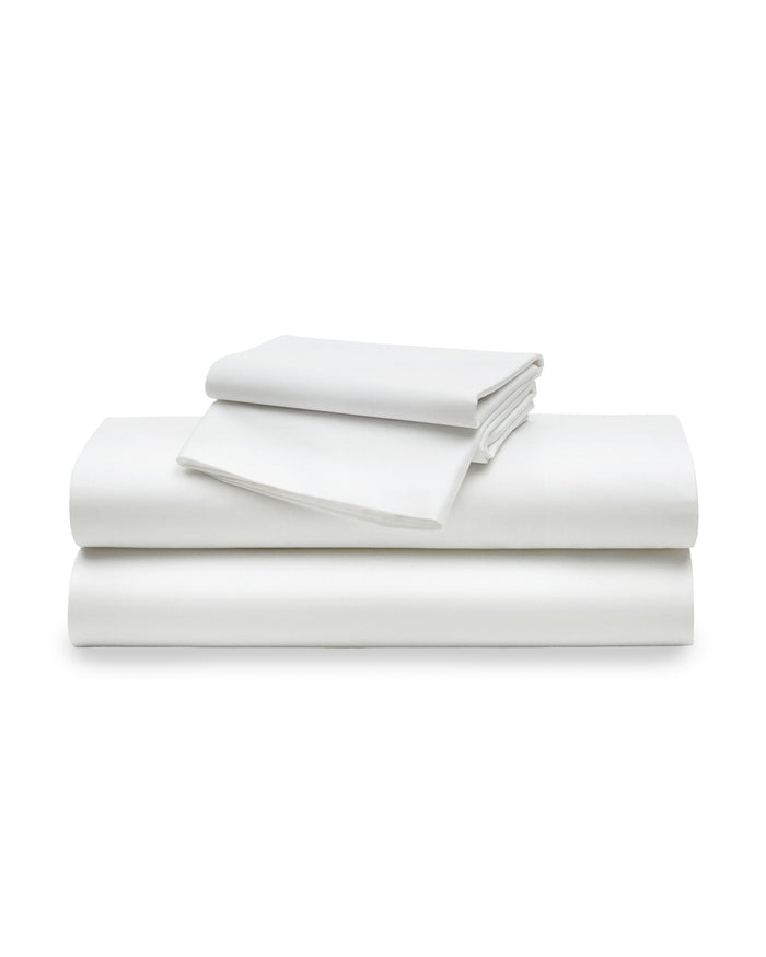 The White Bedding Set includes crisp white sheets, pillowcases, and a comforter, with multiple pillows arranged neatly against a light upholstered headboard in a softly lit bedroom.