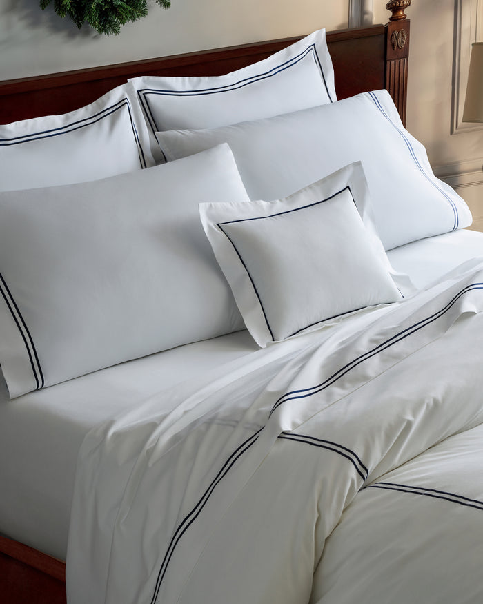A bed with a wooden headboard is dressed in the White With Navy Piping Bedding Set—white sheets, pillows, and a duvet accented by a thin navy border—placed against a light-colored wall.