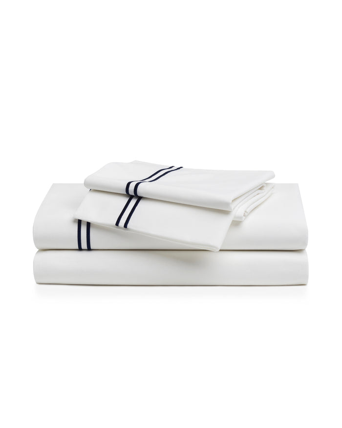A bed with a wooden headboard is dressed in the White With Navy Piping Bedding Set—white sheets, pillows, and a duvet accented by a thin navy border—placed against a light-colored wall.