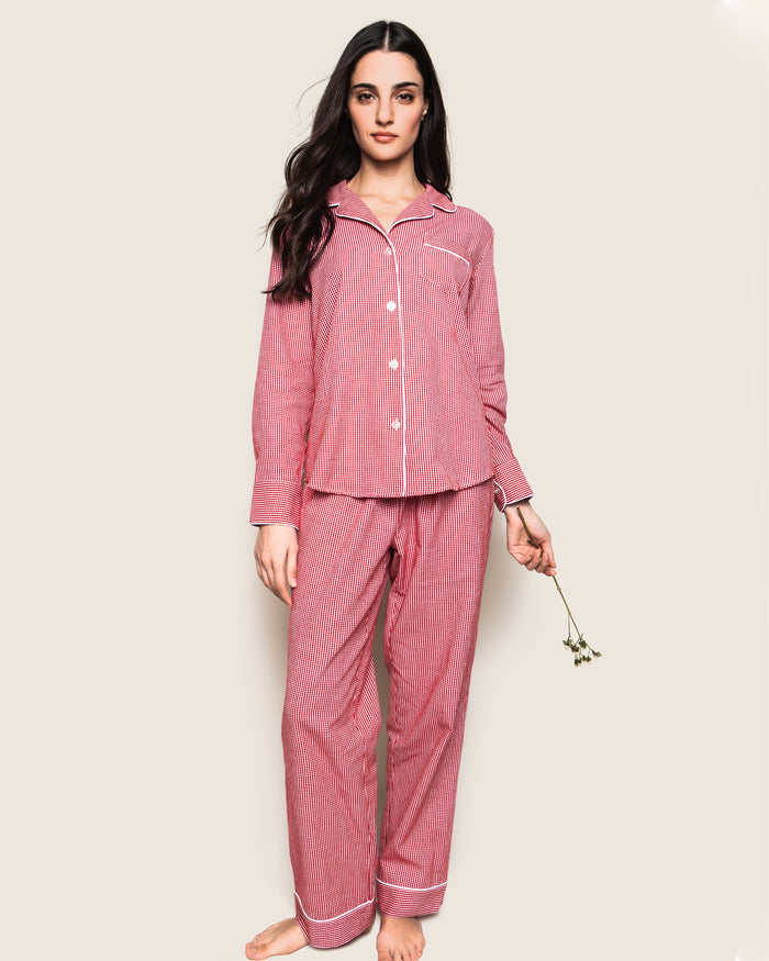 A woman stands barefoot against a plain background, wearing the Womens Flannel Pajama Set in Red Mini Gingham by Petite Plume. She holds small flowers in her right hand, her long dark hair cascading over her shoulders—combining comfort and elegance unique to luxury sleepwear.