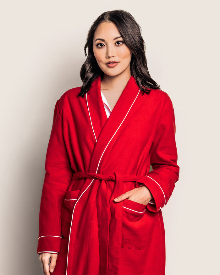 A woman in a Petite Plume Womens Flannel Robe in Red with White Piping, featuring subtle monogramming, stands against a light background. Her shoulder-length dark hair and relaxed expression enhance the cozy vibe, with one hand casually tucked into the robes pocket.
