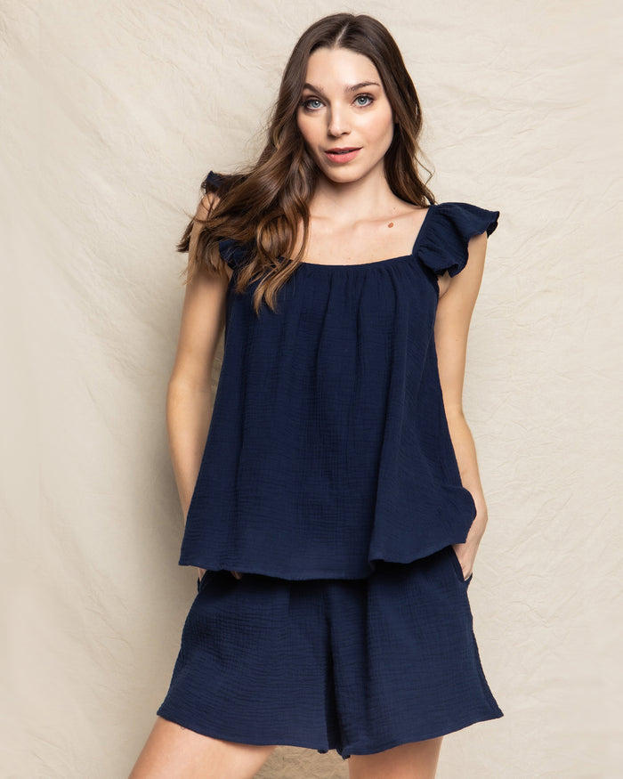 A woman with long hair wears a sleeveless, loose-fitting Womens Gauze Celeste Top in Navy by Petite Plume and matching shorts against a light, textured backdrop. Her hands in pockets, she gazes at the camera with a slight smile, embodying elegant feminine silhouettes.