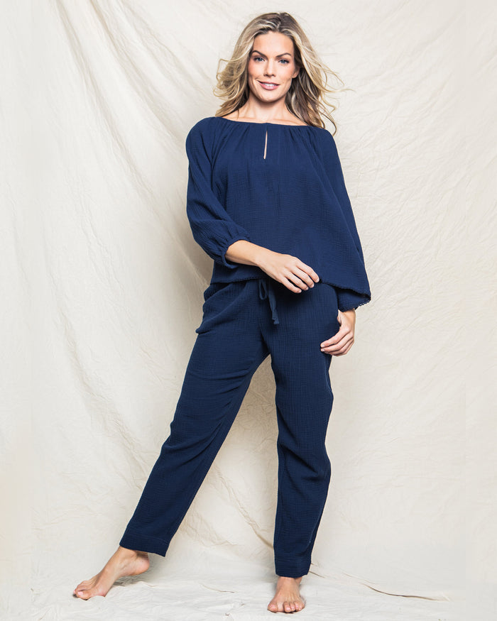 A woman stands barefoot against a light fabric backdrop, wearing Petite Plumes Womens Gauze Willow Scoopneck Top in Navy and matching loose pants. Her wavy hair flows over her shoulders as she smiles, posing with one leg slightly bent.