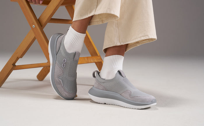A person wearing light gray slip-on sneakers with white crew socks and beige trousers, seated on a wooden folding chair against a neutral studio background | lifestyle