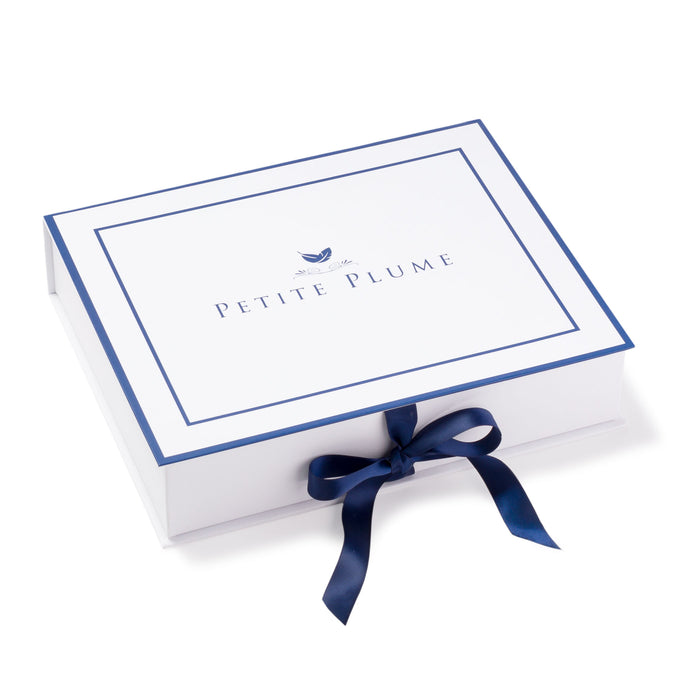 A white gift box, inscribed with Petite Plume in blue and adorned with a feather logo, is elegantly tied with a dark blue ribbon. It hints at the luxe sophistication of the Womens Hamptons Loafer in Black Velvet nestled inside.