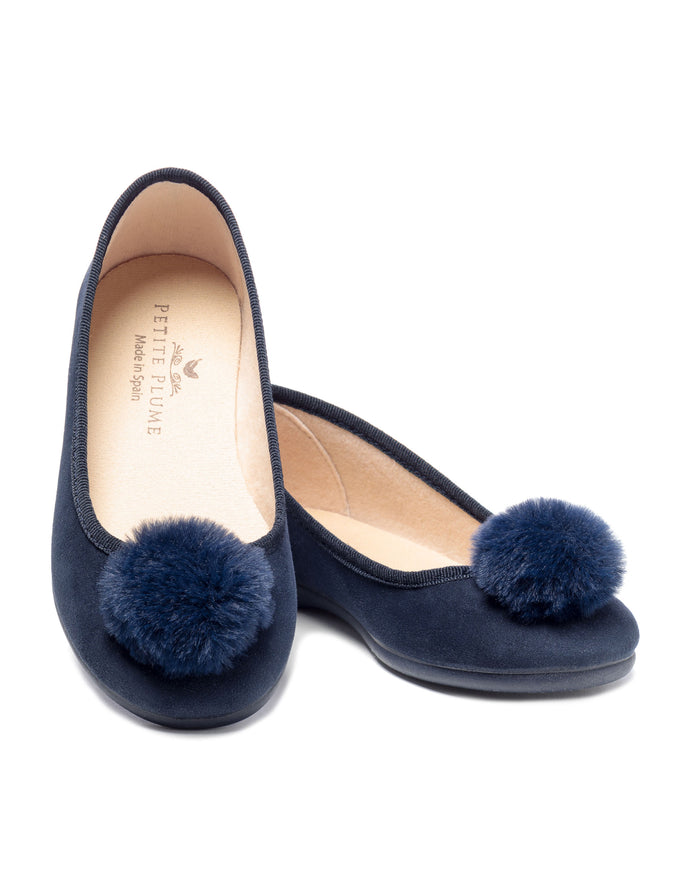 A pair of Petite Plume Womens Juliette Slippers in navy suede with festive pom-poms, showcasing artisanal craftsmanship. They feature a rounded toe, soft beige lining, and are positioned slightly overlapping.