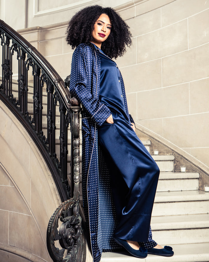 A person with curly hair leans on a staircase railing, wearing a matching dark blue satin outfit and Petite Plumes Womens Juliette Slipper in Navy Suede with a Festive Pom. The elegant setting, with its grand staircase and ornate railing, exudes sophisticated artisanal craftsmanship.