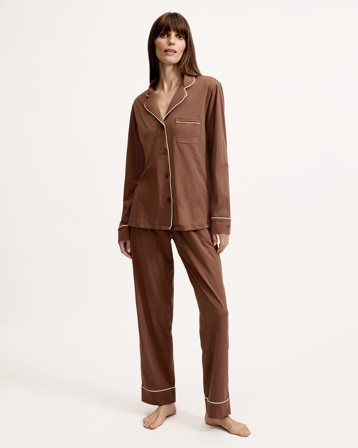 A woman stands barefoot in the Women's Lightweight Pima Pajama Set in Chocolate Brown, featuring a button-up long-sleeve shirt with white piping and matching pants, against a plain white background.