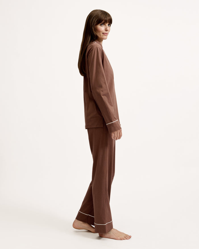 A woman stands barefoot in the Women's Lightweight Pima Pajama Set in Chocolate Brown, featuring a button-up long-sleeve shirt with white piping and matching pants, against a plain white background.