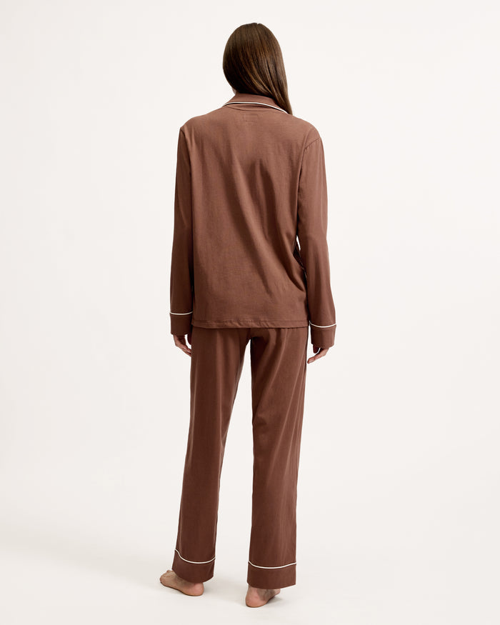 A woman stands barefoot in the Women's Lightweight Pima Pajama Set in Chocolate Brown, featuring a button-up long-sleeve shirt with white piping and matching pants, against a plain white background.