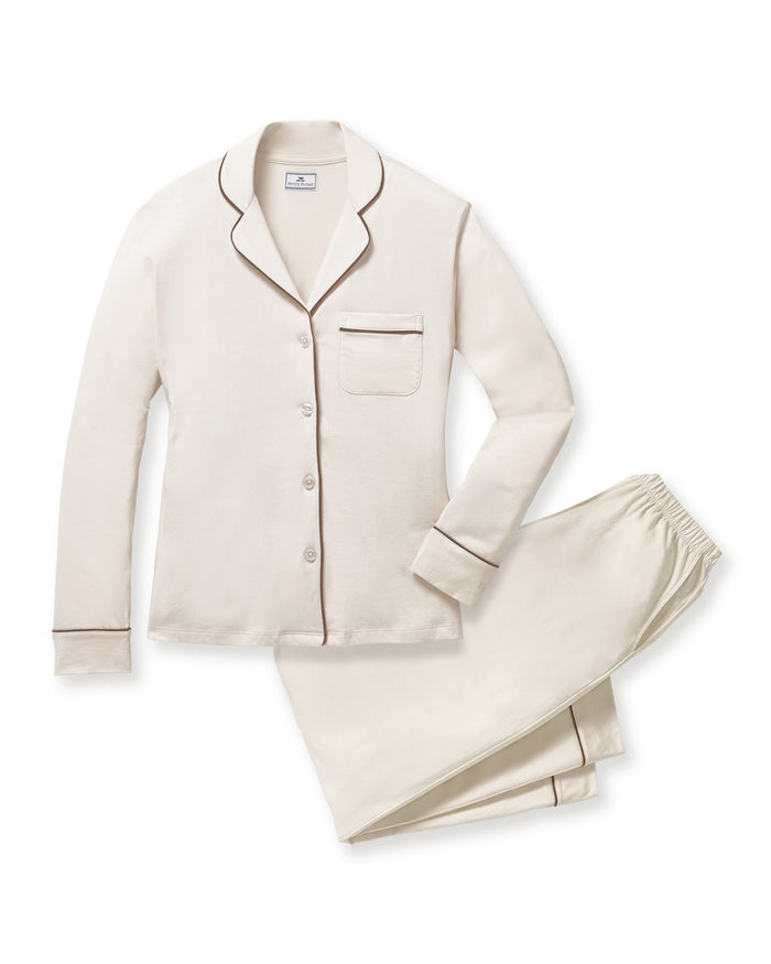 A woman stands barefoot against a plain background, wearing the Women's Lightweight Pima Pajama Set in Ivory featuring dark piping, a long-sleeve button-up top, and matching long pants.