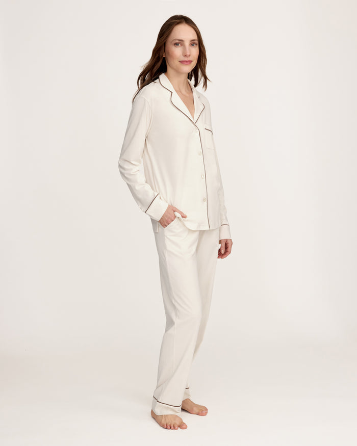 A woman stands barefoot against a plain background, wearing the Women's Lightweight Pima Pajama Set in Ivory featuring dark piping, a long-sleeve button-up top, and matching long pants.