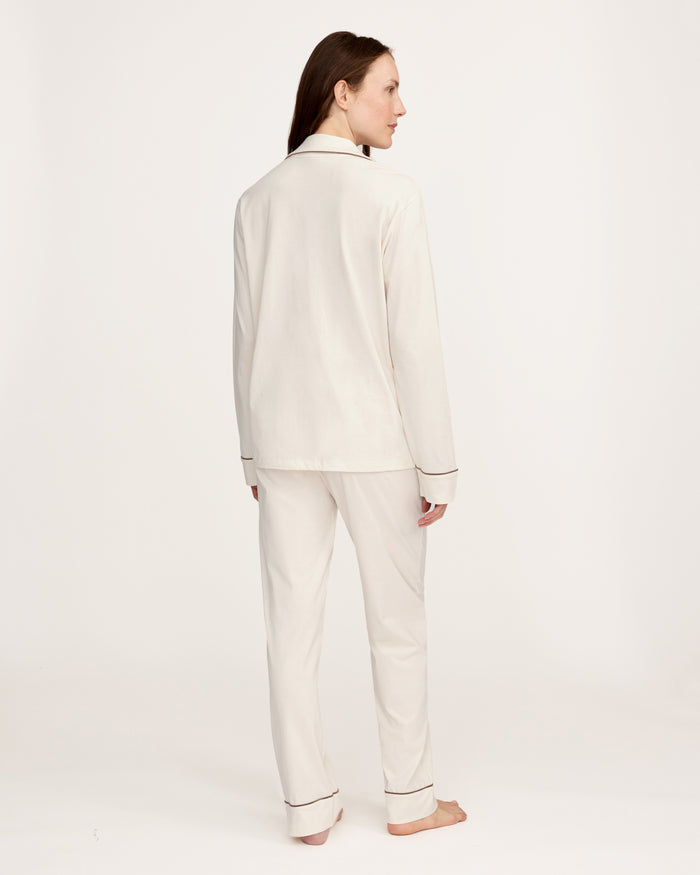 A woman stands barefoot against a plain background, wearing the Women's Lightweight Pima Pajama Set in Ivory featuring dark piping, a long-sleeve button-up top, and matching long pants.