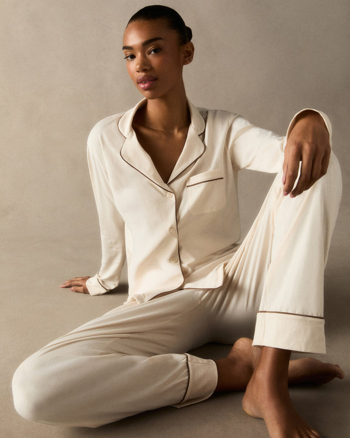 A woman stands barefoot against a plain background, wearing the Women's Lightweight Pima Pajama Set in Ivory featuring dark piping, a long-sleeve button-up top, and matching long pants.