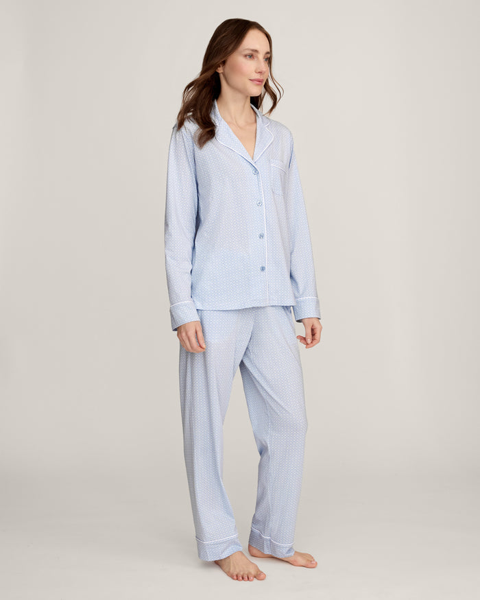 A woman with light brown hair and a neutral expression stands barefoot against a plain background, modeling the Women's Lightweight Pima Pajama Set in Periwinkle Geo—featuring long sleeves, button-up top with collar, and matching pants.
