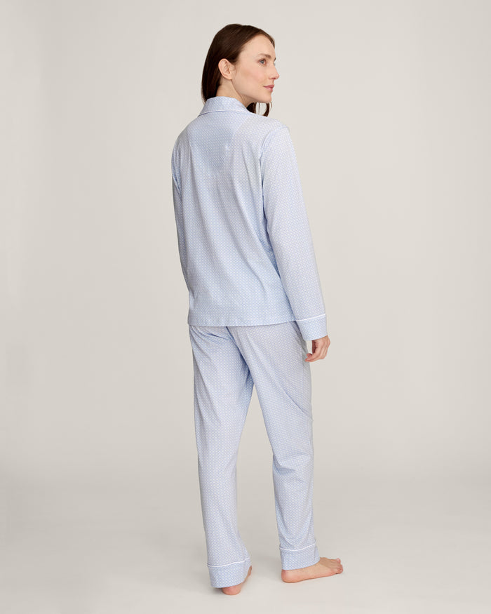A woman with light brown hair and a neutral expression stands barefoot against a plain background, modeling the Women's Lightweight Pima Pajama Set in Periwinkle Geo—featuring long sleeves, button-up top with collar, and matching pants.