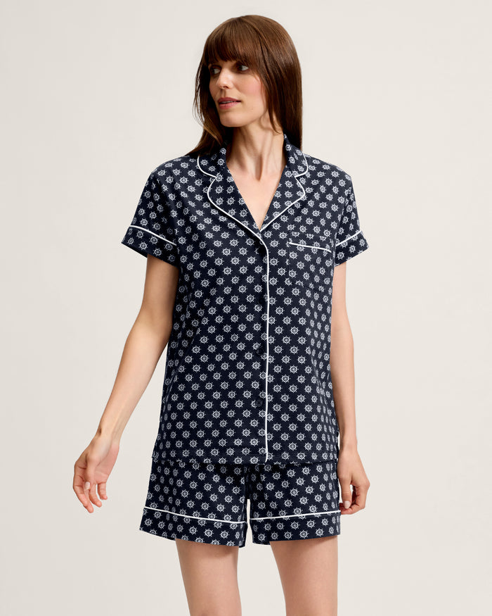 A woman wearing the Women’s Lightweight Pima Short Sleeve Short Set in Captain’s Compass, featuring a navy blue fabric with white geometric patterns and white piping, stands against a plain light background, gazing to the side.