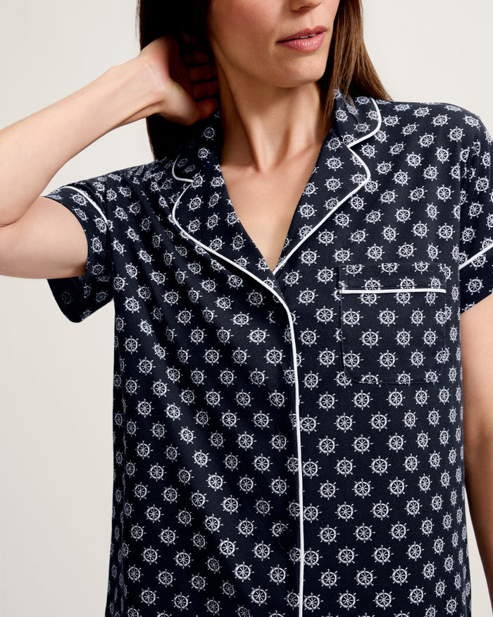 A woman wears the Women’s Lightweight Pima Short Sleeve Short Set in Captain’s Compass—navy blue pajamas adorned with white nautical ship wheel patterns and finished with white piping. She stands with one hand behind her neck, her face partially out of frame.