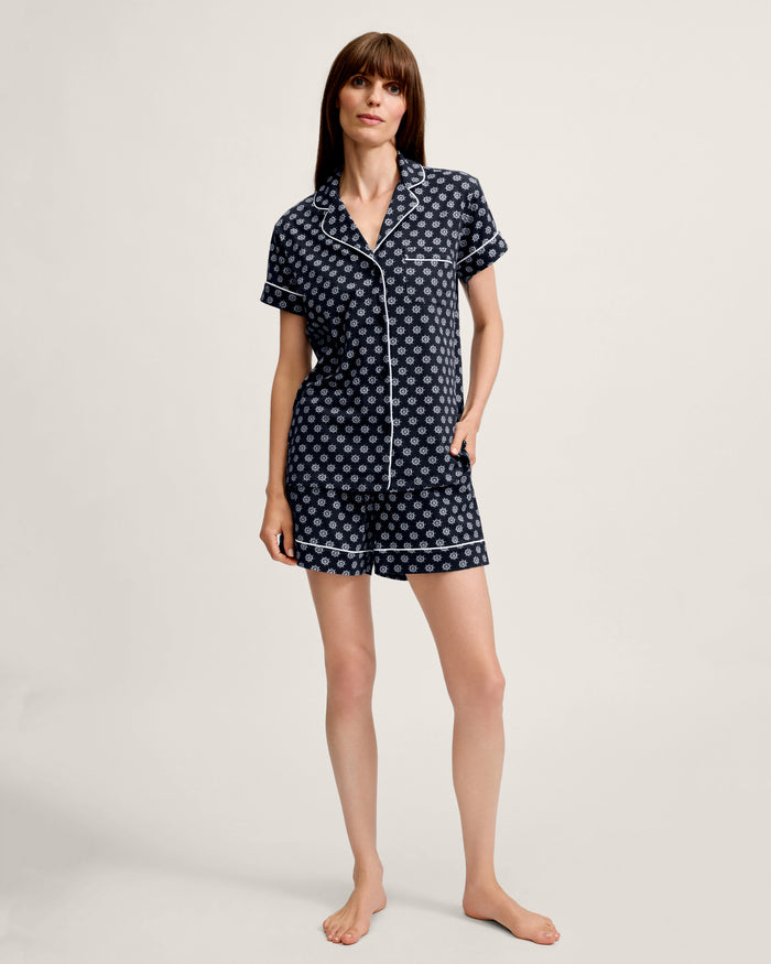A woman stands barefoot against a plain backdrop, wearing the Women’s Lightweight Pima Short Sleeve Short Set in Captain’s Compass, which features a subtle white pattern and white trim; her left hand rests in her shorts pocket while her right arm hangs relaxed at her side.