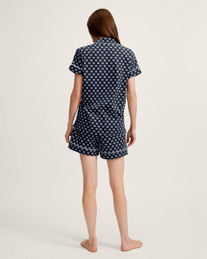 A woman with long hair stands barefoot, facing away, wearing the Women’s Lightweight Pima Short Sleeve Short Set in Captain’s Compass—a matching short-sleeve shirt and shorts featuring a small white geometric pattern on a dark background. The setting is a plain, light-colored backdrop.