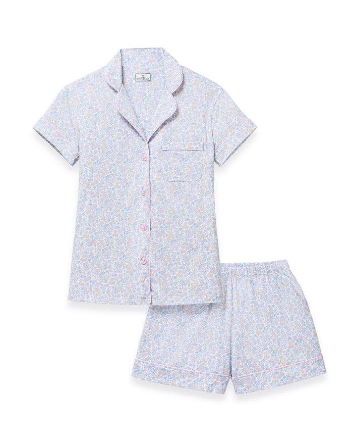 A woman with long wavy hair wears the Women's Lightweight Pima Short Sleeve Short Set in Fleur Blanche, featuring a pastel floral pattern, standing against a plain background.