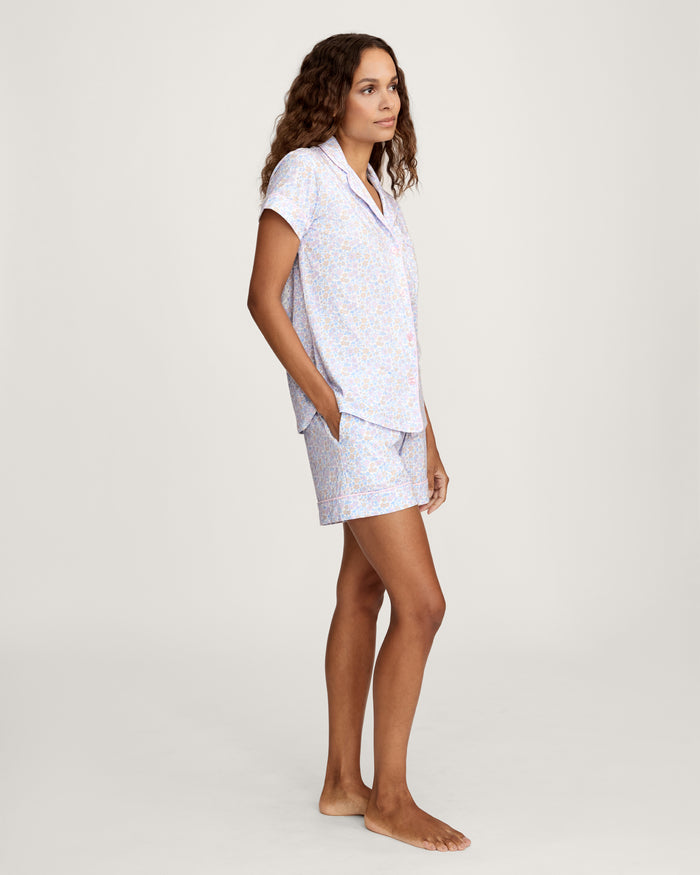 A woman with long wavy hair wears the Women's Lightweight Pima Short Sleeve Short Set in Fleur Blanche, featuring a pastel floral pattern, standing against a plain background.