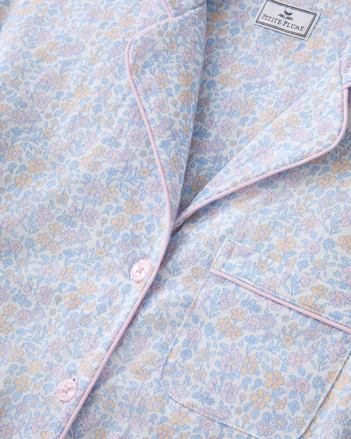 A woman with long wavy hair wears the Women's Lightweight Pima Short Sleeve Short Set in Fleur Blanche, featuring a pastel floral pattern, standing against a plain background.
