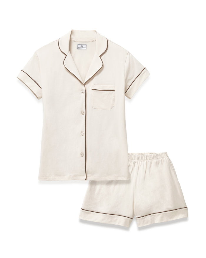 A woman with long curly hair models the Women's Lightweight Pima Short Sleeve Short Set in Ivory, featuring a button-up top with piping and matching shorts, standing barefoot against a plain light background.