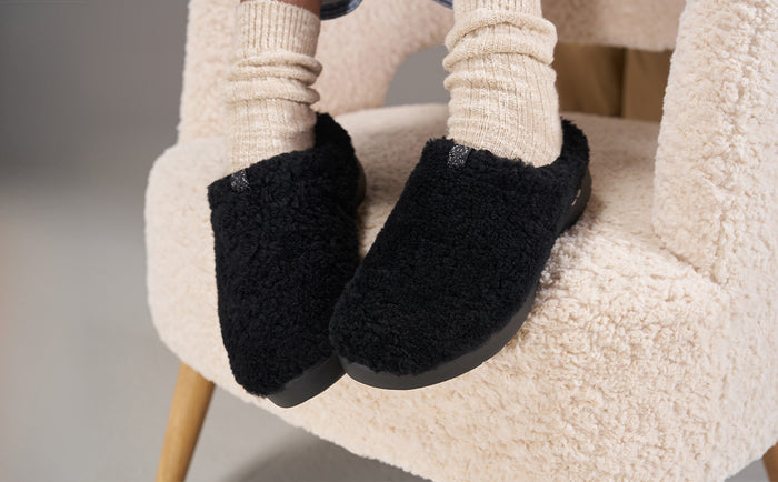 A close-up of a person sitting on a cream-colored sheepskin chair, wearing black fuzzy slip-on slippers and cream-colored socks | lifestyle