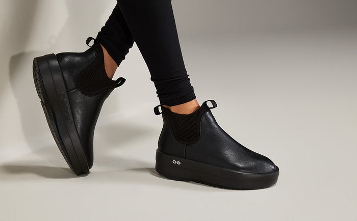 A person wearing black leather Chelsea-style platform boots with black elastic side panels and black leggings, photographed against a clean light gray studio background | lifestyle