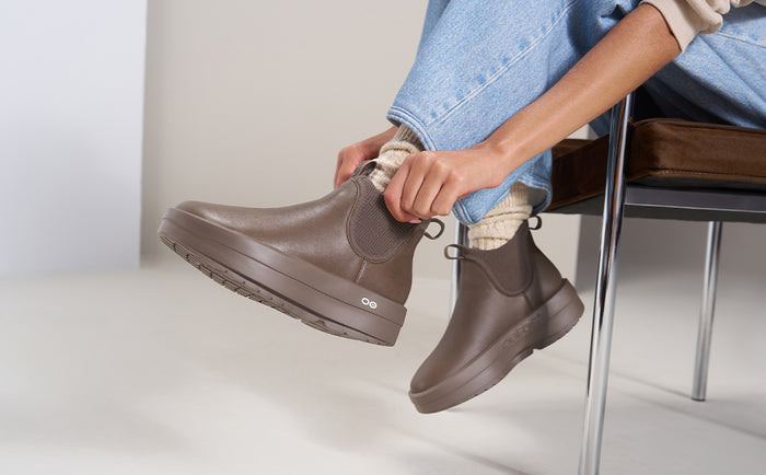 A person sitting on a chair wearing taupe-brown rubber Chelsea-style boots with elastic side panels and chunky soles, paired with light blue jeans and cream socks | lifestyle