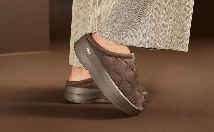 A person wearing brown quilted slip-on mule shoes with chunky gray soles, shown from the calves down against a warm brown studio background | lifestyle