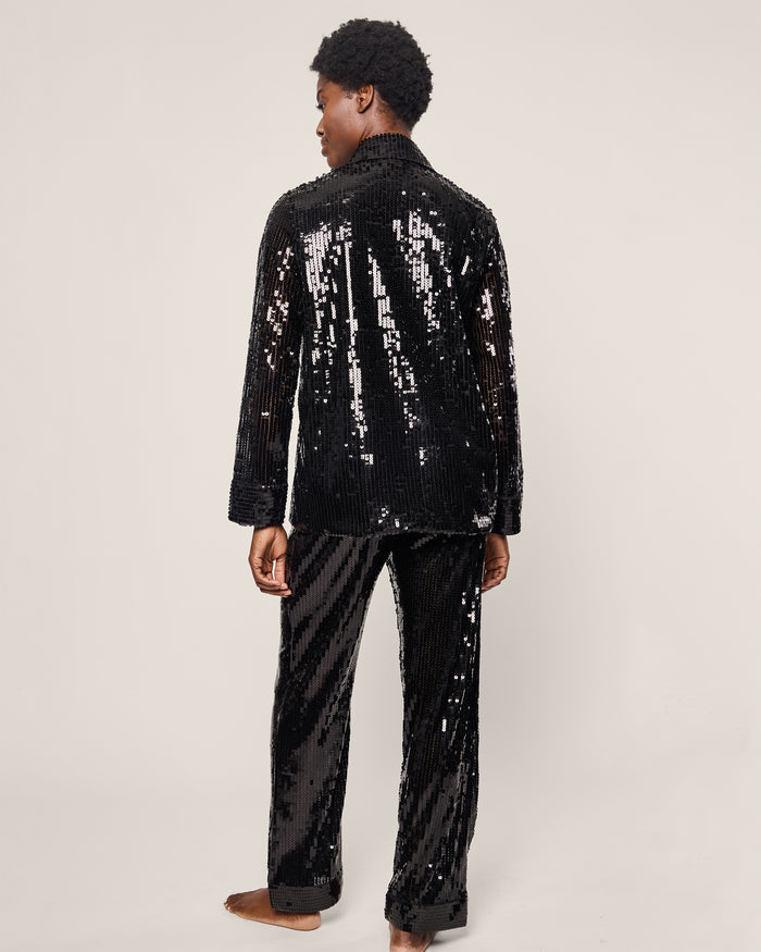 A person in Petite Plumes Womens Pajama Set in Black Sequins stands barefoot against a plain backdrop, slightly turning away from the camera. The reflective long sleeves and full-length pants blend luxury sleepwear with captivating style.
