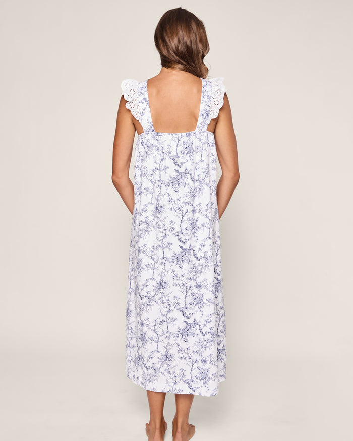 A long-haired person is depicted from the back wearing Petite Plumes Womens Pima Clara Nightgown in Timeless Toile. The sleeveless, white and blue floral dress with lace shoulder details drapes elegantly to ankle-length against a light gray background, evoking a serene bedtime ritual.