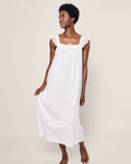 Against a plain backdrop, a woman stands barefoot in the Womens Pima Clara Nightgown in White by Petite Plume. This sleeveless gown with lace shoulder details resembles elegant jersey knit pajamas. She looks aside with a relaxed expression, gently holding her dress.