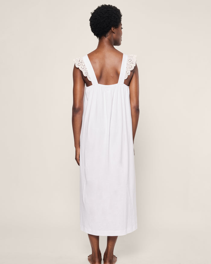 A person with short hair, barefoot and facing away, wears the Womens Pima Clara Nightgown by Petite Plume. The sleeveless white gown features Peruvian Pima cotton with lace-detailed straps against a light beige backdrop, evoking a calming bedtime ambiance.