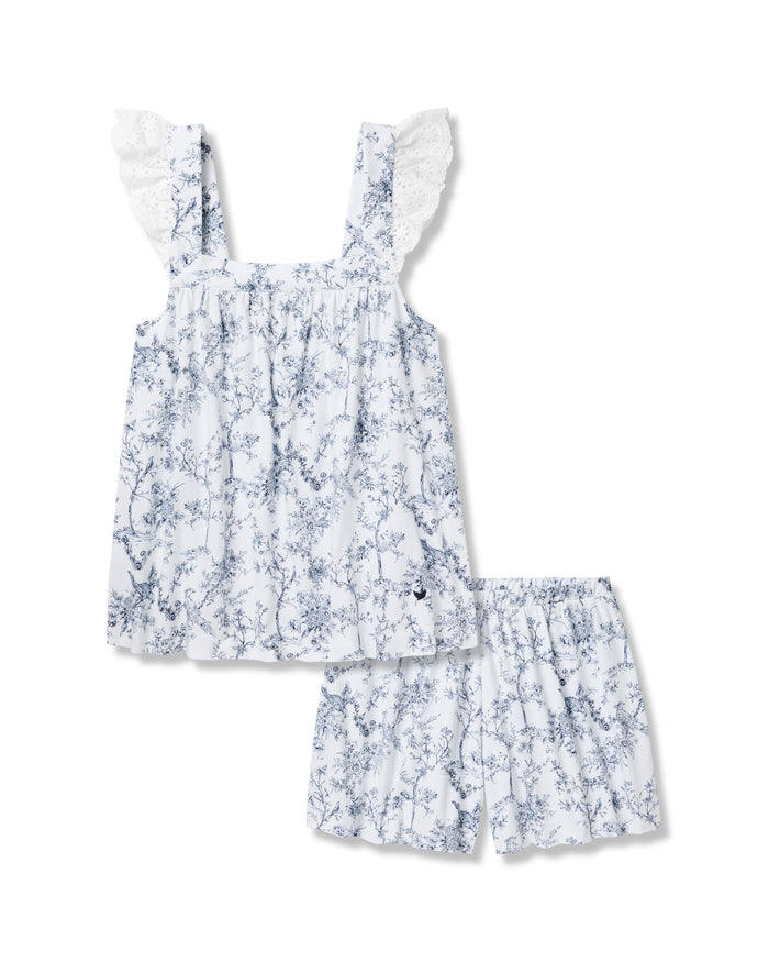 Wearing the Womens Pima Clara Short Set in Timeless Toile by Petite Plume, featuring a blue floral pattern and ruffled shoulder straps, one exudes botanical elegance with their short curly hair against a plain background.