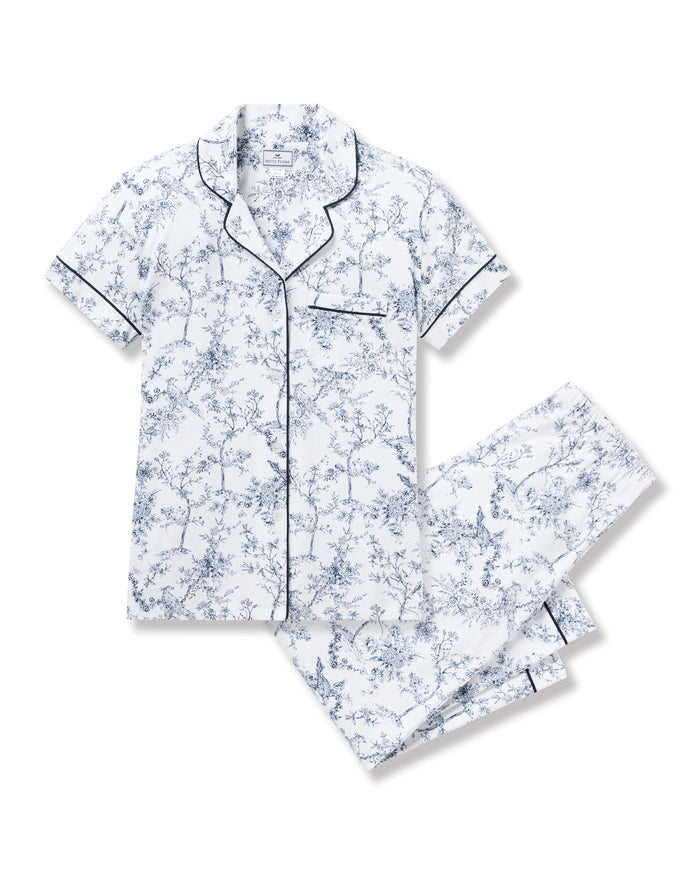A woman embodies botanical elegance in the Womens Pima Crop Set in Timeless Toile by Petite Plume, featuring a matching pajama set with a blue floral pattern. The short-sleeve collared top pairs with loose-fitting pants as she stands barefoot against a plain light background.