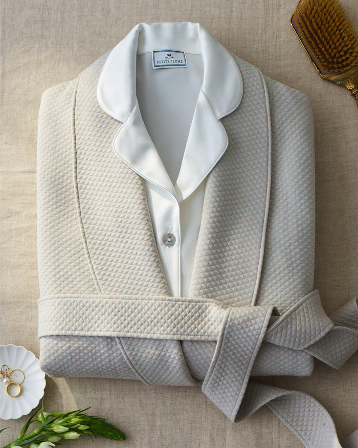 The Women’s Pima Jacquard Ophelia Robe in Taupe, featuring a textured pattern and matching belt, is neatly folded on a neutral cloth. A white-collared pajama top peeks out at the neckline, while a gold ring dish, green leaves, and a wooden hairbrush are arranged nearby.