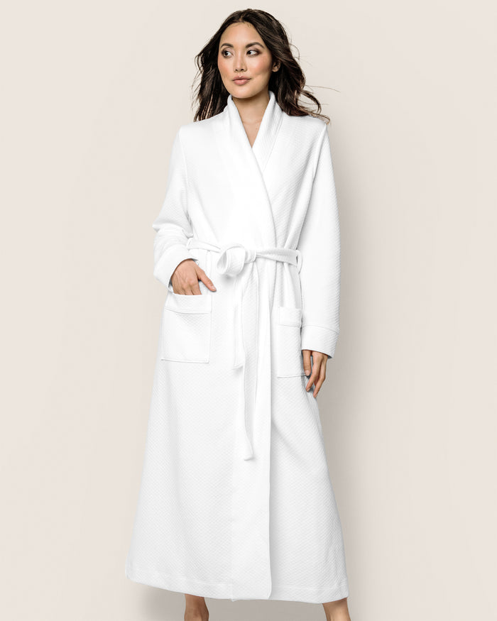 Wearing the Womens Pima Ophelia Robe in White Jacquard by Petite Plume, they stand poised against a plain backdrop. The luxurious Peruvian Pima cotton robe features an elegant quilted texture, two front pockets, and a waist belt. Long, dark hair flows down their back gracefully.